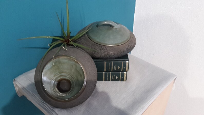 May include: Two ceramic vessels with a stone-like texture, one open and the other with a lid. The open vessel holds a small plant. Both have a green interior and sit on a stack of books and a linen cloth.