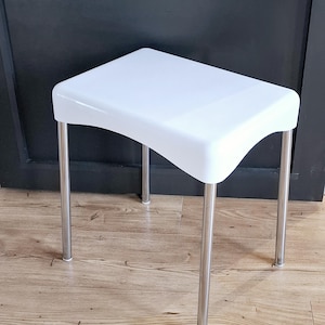 May include: White plastic stool with chrome legs. The stool has a slightly curved top.