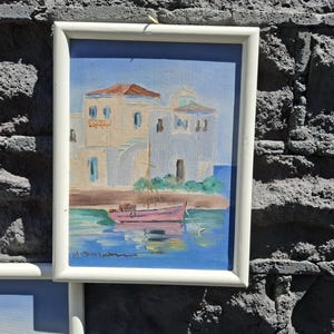 May include: A framed painting depicting a coastal scene with white buildings featuring a dome, a pink boat floating on blue water, and a clear sky. The artwork is enclosed in a simple white rectangular frame, showcasing a Mediterranean-style landscape.