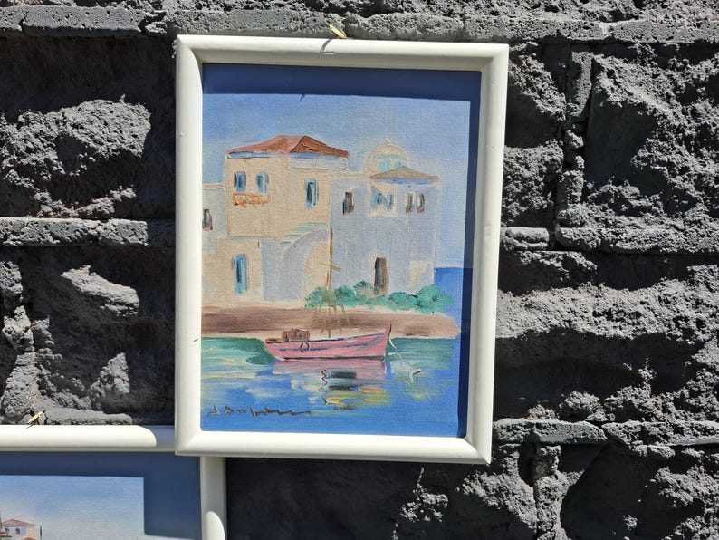 May include: A framed painting depicting a coastal scene with white buildings featuring a dome, a pink boat floating on blue water, and a clear sky. The artwork is enclosed in a simple white rectangular frame, showcasing a Mediterranean-style landscape.