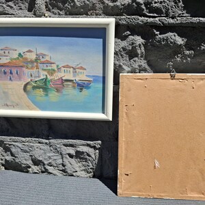 May include: A framed landscape painting depicting a coastal village with white buildings, red roofs, and colorful boats floating in blue water. Beside it is a second rectangular frame shown from the back, featuring a plain brown cardboard backing.