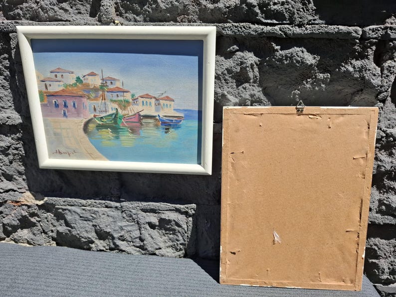 May include: A framed landscape painting depicting a coastal village with white buildings, red roofs, and colorful boats floating in blue water. Beside it is a second rectangular frame shown from the back, featuring a plain brown cardboard backing.