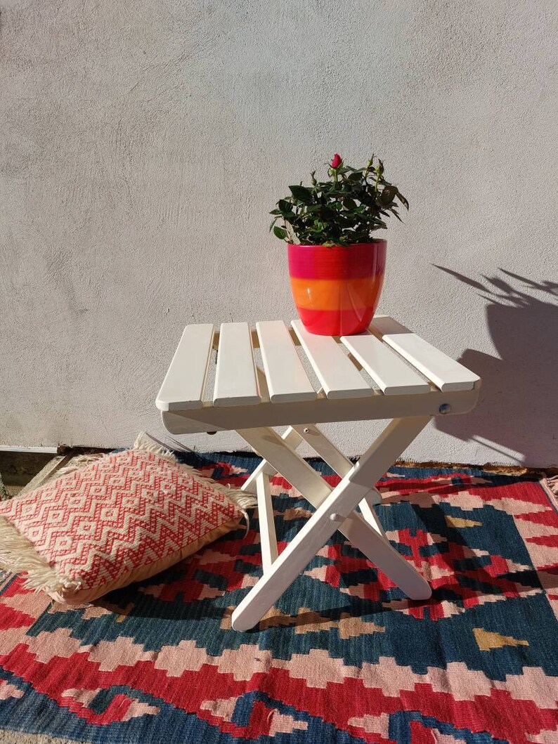 May include: A white folding table with a potted plant on top. The table is on a colorful rug with a red, white, and blue pattern. The plant is in a red, orange, and yellow pot.