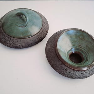 Pode incluir: Dois vasos de cerâmica com esmalte verde-azulado e bordas escuras e texturizadas. Um vaso tem uma tampa com uma alça em espiral. O outro vaso está aberto, revelando um interior profundo. A cerâmica tem uma aparência rústica e artesanal.