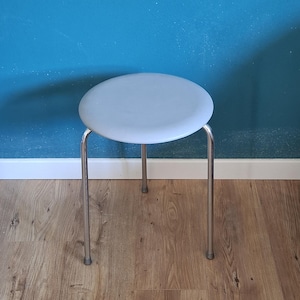 Può includere: Uno sgabello rotondo grigio chiaro con struttura in metallo. Lo sgabello ha un design semplice con quattro gambe e un sedile circolare. Le gambe sono in metallo lucido e il sedile è di colore grigio chiaro uniforme. Lo sgabello è appoggiato a un muro blu.
