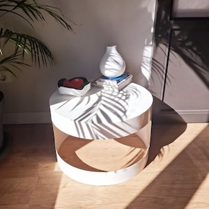 Op de afbeelding: Een witte, ronde bijzettafel met een onderste plank. Een witte vaas, een kleine decoratieve kom met een rode binnenkant en boeken staan erop. De tafel staat in een kamer met houten vloeren en een palmboom.
