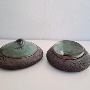 May include: Two ceramic decorative bowls. One has a lid with a handle, the other is open. Both have a dark, textured base and a teal-colored interior. The lid has a teal handle. The bowls are on a white surface.