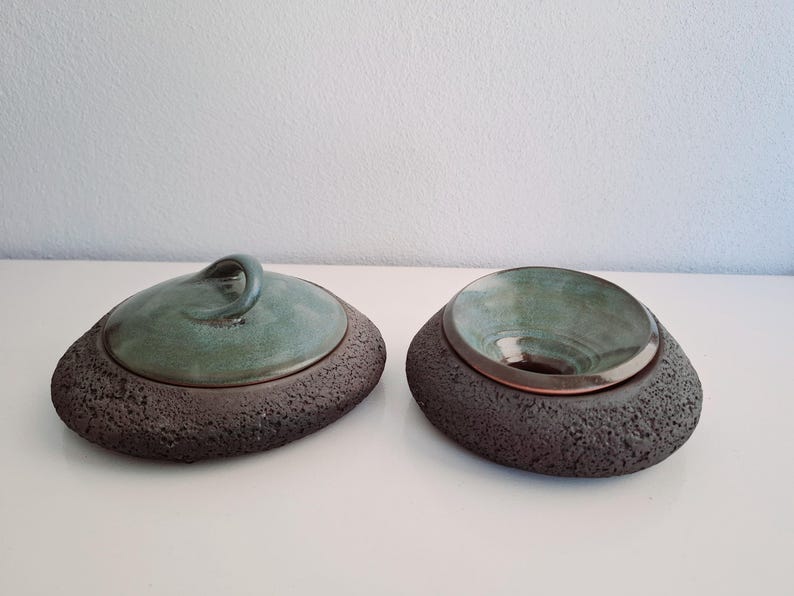 May include: Two ceramic decorative bowls. One has a lid with a handle, the other is open. Both have a dark, textured base and a teal-colored interior. The lid has a teal handle. The bowls are on a white surface.