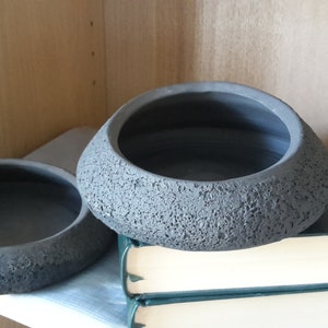 May include: Two gray, textured ceramic bowls with a dark interior. The bowls have a unique, organic shape and sit on a stack of books. The bowls are displayed on a shelf, suggesting a decorative or functional use.