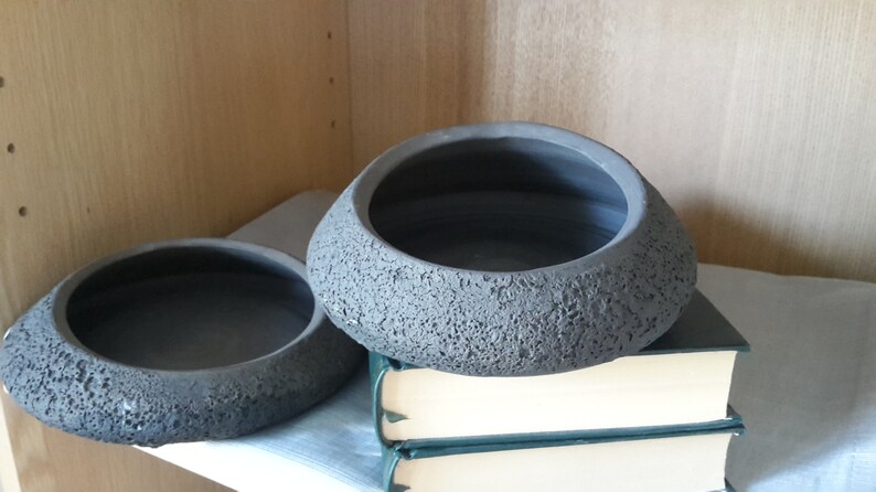 May include: Two gray, textured ceramic bowls with a dark interior. The bowls have a unique, organic shape and sit on a stack of books. The bowls are displayed on a shelf, suggesting a decorative or functional use.
