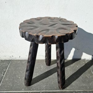 May include: A dark brown, three-legged wooden stool. The round seat has a scalloped edge and carved floral pattern. The legs are also carved with a repeating design. The stool is likely handmade and has a rustic, vintage aesthetic.