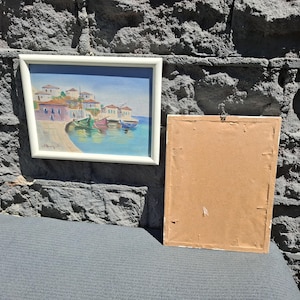 May include: A framed landscape painting depicting a coastal village with white buildings, red roofs, and colorful boats docked in blue water, positioned next to a second rectangular frame showing only its plain brown cardboard backing.
