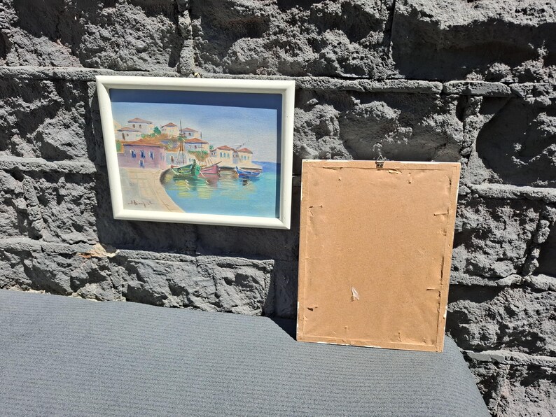 May include: A framed landscape painting depicting a coastal village with white buildings, red roofs, and colorful boats docked in blue water, positioned next to a second rectangular frame showing only its plain brown cardboard backing.
