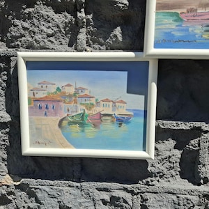 May include: A framed painting depicting a coastal village with white buildings and red roofs situated along a harbor. Several small colorful boats are docked in the turquoise water. The artwork features a signature in the bottom left corner.