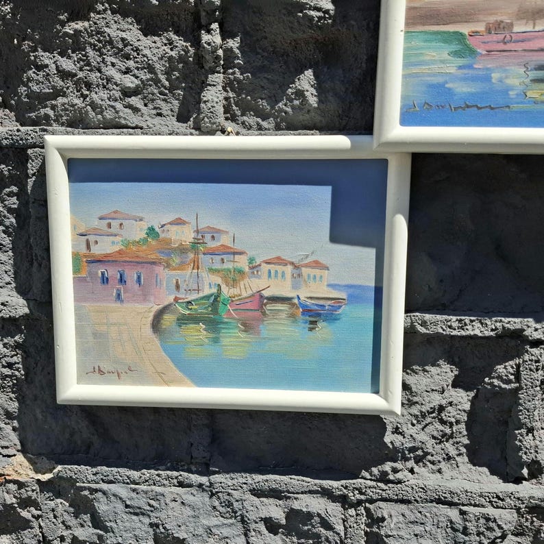 May include: A framed painting depicting a coastal village with white buildings and red roofs situated along a harbor. Several small colorful boats are docked in the turquoise water. The artwork features a signature in the bottom left corner.