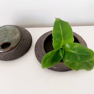 May include: Two dark brown ceramic bowls with textured exteriors. One bowl has a teal interior and a lid. The other bowl contains green leaves. The bowls are on a white surface.