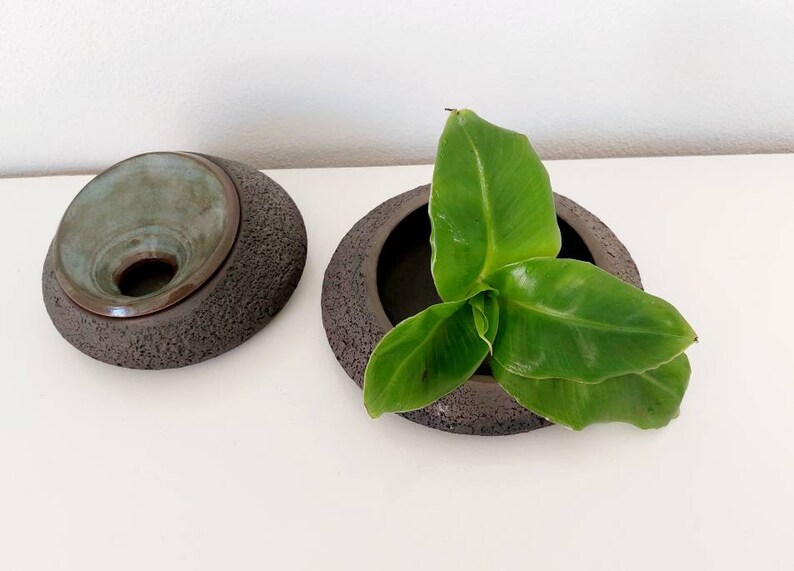 May include: Two dark brown ceramic bowls with textured exteriors. One bowl has a teal interior and a lid. The other bowl contains green leaves. The bowls are on a white surface.