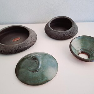 May include: Four handcrafted ceramic pieces in shades of gray and teal. The collection includes two open bowls, a lid with a curved handle, and a small, flared bowl with a central hole. The bowls have textured and smooth surfaces.