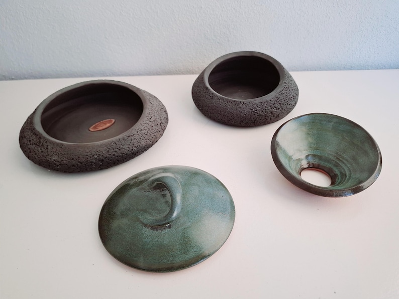 May include: Four handcrafted ceramic pieces in shades of gray and teal. The collection includes two open bowls, a lid with a curved handle, and a small, flared bowl with a central hole. The bowls have textured and smooth surfaces.