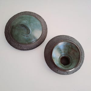 May include: Two ceramic bowls with a textured, dark gray rim and a teal interior. One bowl has a raised, curved design, while the other has a conical shape. The bowls are displayed against a white background.