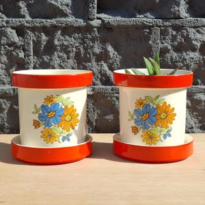 Pode incluir: Dois vasos cilíndricos de cerâmica com pratos a condizer, apresentando um corpo de cor creme e um rebordo laranja vibrante. Cada vaso está decorado com um padrão floral de flores azuis, cor de laranja e amarelas com folhas verdes.