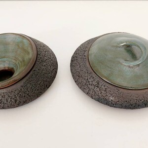 May include: Two ceramic art pieces. Each has a dark, textured base with a teal-colored, bowl-shaped top. One has an open bowl, the other has a lid with a swirl handle. The pieces are on a white surface.