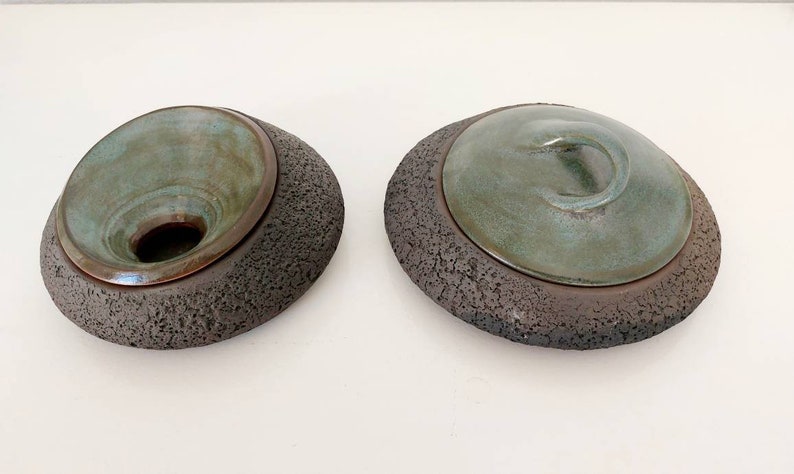 May include: Two ceramic art pieces. Each has a dark, textured base with a teal-colored, bowl-shaped top. One has an open bowl, the other has a lid with a swirl handle. The pieces are on a white surface.