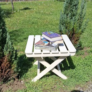 May include: A white folding stool with three books stacked on top. The books are a purple book, a blue book, and a brown book. The stool is sitting on a green grassy lawn.