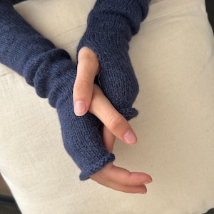 Op de afbeelding: Een paar marineblauwe vingerloze handschoenen. De gebreide handschoenen hebben een geribbelde manchet en een duimgat. De handschoenen zijn gemaakt van een zacht, getextureerd materiaal. De handschoenen worden getoond op een lichtbeige oppervlak.