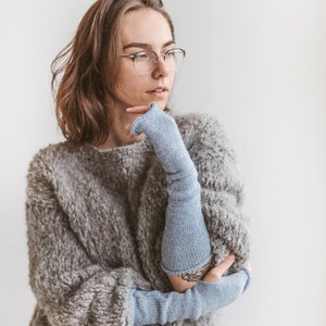 Può includere: Una donna indossa un maglione grigio peloso e guanti senza dita di colore blu chiaro. Indossa occhiali e guarda da un'altra parte.
