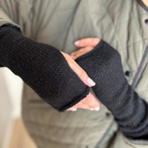 Peut inclure: Une paire de mitaines noires en tricot. Les gants sont présentés sur un fond de veste matelassée vert olive. Les gants couvrent la main jusqu'aux jointures, offrant chaleur et dextérité.