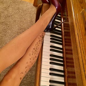 May include: A pair of burgundy high heels with a stiletto heel, worn by a person with a tattoo of musical notes and dandelions on their legs. The person is sitting on a wooden piano with the keys visible.