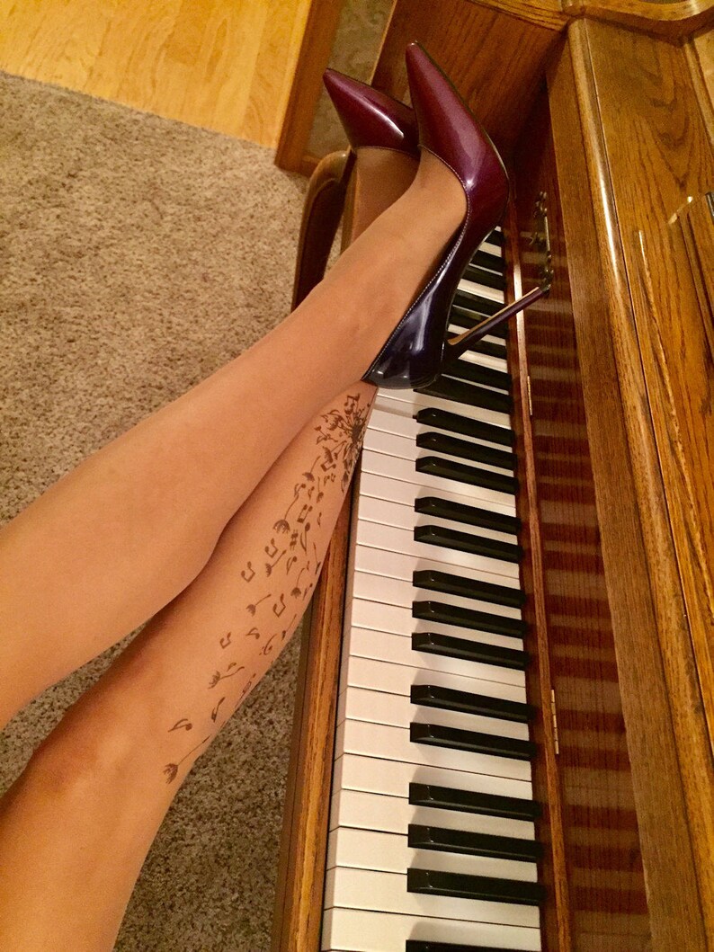 May include: A pair of burgundy high heels with a stiletto heel, worn by a person with a tattoo of musical notes and dandelions on their legs. The person is sitting on a wooden piano with the keys visible.