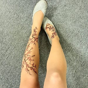 Puede incluir: Un par de medias de color carne con un dise&ntilde;o floral en las piernas. El dise&ntilde;o presenta una rama de cerezos en flor con flores rosas y tallos marrones oscuros. La persona lleva zapatos planos grises.