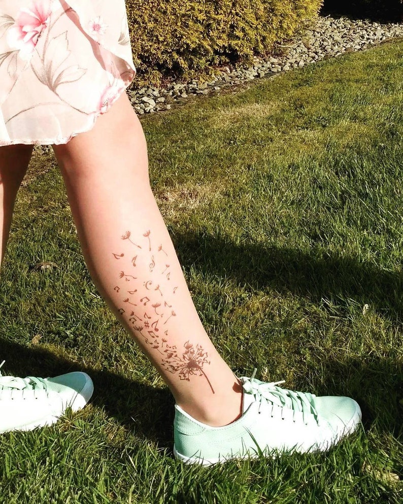 May include: A person's leg with a dandelion tattoo on the lower calf. The tattoo features a dandelion with musical notes blowing in the wind. The person is wearing a light green pair of sneakers.