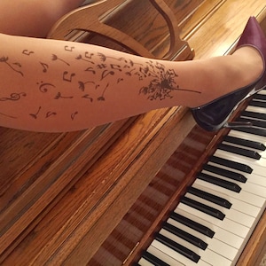 May include: A woman's leg with a tattoo of musical notes and a dandelion on it. She is wearing a pair of dark purple high heels and is sitting at a piano.