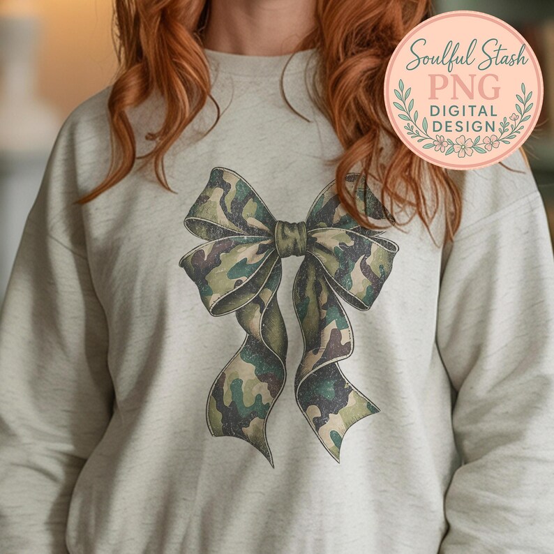 May include: A light gray sweatshirt featuring a large bow design in a camouflage pattern of green, brown, and tan. The bow is centered on the front of the shirt. The image also includes a circular logo with the text "Soulful Stash PNG DIGITAL DESIGN".