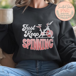 May include: Dark grey sweatshirt with the slogan "Just Keep Spinning" in white and red, featuring a graphic of a stationary exercise bike. The design is vintage style, suitable for fitness enthusiasts.