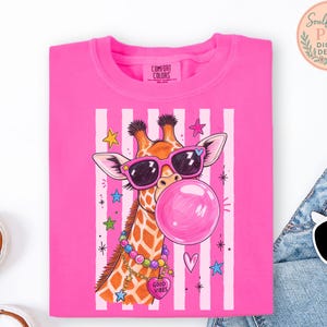 A pink t-shirt with a giraffe wearing sunglasses and a bubble gum bubble, surrounded by colorful stars and hearts.