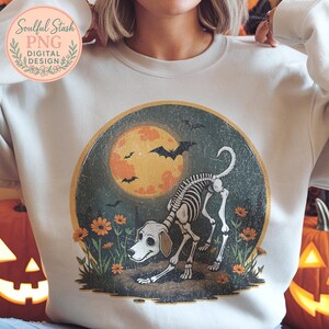 May include: Cream-colored sweatshirt featuring a Halloween-themed graphic. The design includes a dog skeleton, a full moon, bats, and flowers. The text "Soulful Stash PNG DIGITAL DESIGN" is in the upper left corner.