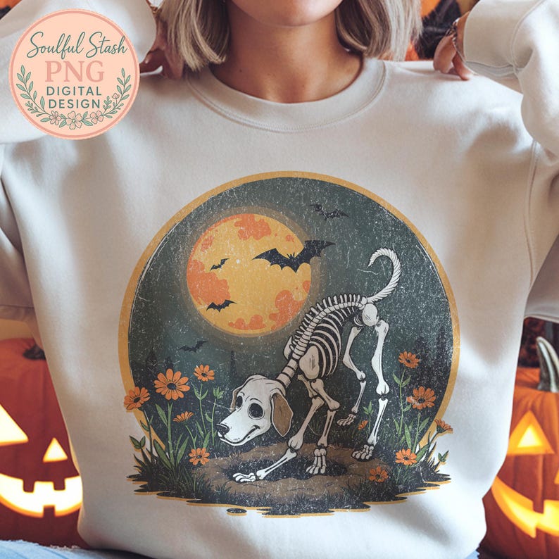 May include: Cream-colored sweatshirt featuring a Halloween-themed graphic. The design includes a dog skeleton, a full moon, bats, and flowers. The text "Soulful Stash PNG DIGITAL DESIGN" is in the upper left corner.