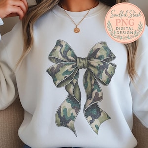 May include: White sweatshirt featuring a large bow design in a camouflage pattern. The bow is predominantly green, brown, and tan, with detailed shading. The sweatshirt has a crew neck and long sleeves. A circular logo with text is visible in the upper right corner.