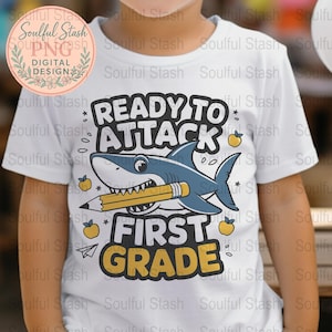 May include: White t-shirt featuring a cartoon shark holding a pencil, with the text "READY TO ATTACK FIRST GRADE". The shark is blue and white, with yellow accents. The shirt is for children.