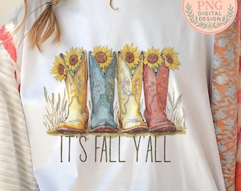May include: White t-shirt featuring a watercolor illustration of four colorful cowboy boots, each holding a sunflower. The text "IT'S FALL Y'ALL" is printed below the boots. A digital design logo is in the upper right corner.