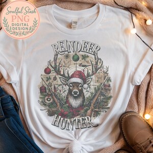 May include: White t-shirt featuring a detailed graphic of a reindeer wearing a Santa hat, antlers adorned with Christmas lights, and crossed rifles. The text "REINDEER HUNTER" is displayed above and below the reindeer illustration.