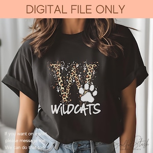 May include: A black t-shirt with a white paw print and a leopard print letter W. The text "WILDCATS" is printed below the letter. The shirt is being modeled by a person.