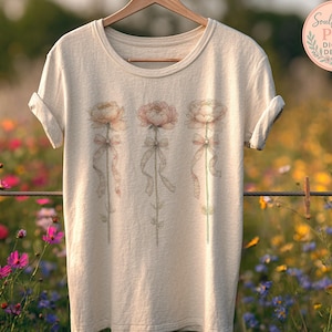 Puede incluir: Una camiseta color crema con tres ilustraciones de flores de peonía estilo acuarela con cintas. Las flores son en tonos rosa y melocotón, con tallos verdes. La camiseta está colgada en una percha de madera.