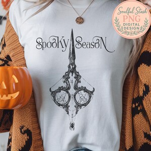 May include: White t-shirt with a black graphic of ornate scissors, spiderwebs, and a spider. The words "Spooky Season" are above the scissors. A carved pumpkin and a floral logo are also visible.