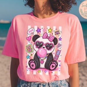 A woman wearing a pink t-shirt with a colorful graphic of a panda wearing sunglasses and a bow, surrounded by various decorative elements.