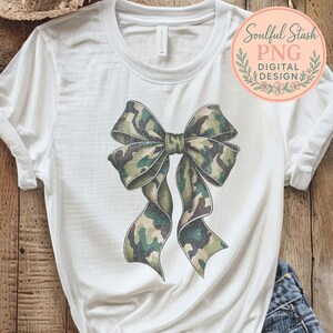 May include: White t-shirt featuring a large bow design in a camouflage pattern of green, brown, and tan. The bow is the focal point, with detailed shading. The shirt also has the text "Soulful Stash PNG Digital Design" in a circular logo.